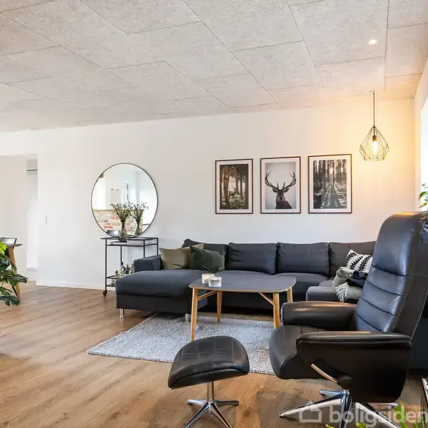 En stue med en mørkegrå sofa står mod en hvid mur med billeder af en hjort og skove. Foran sofaen er et lille bord med en lampe hængende over det.