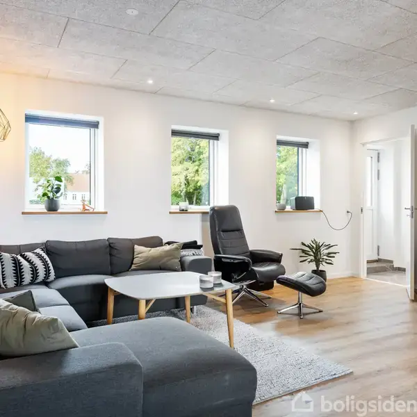 Stor, grå hjørnesofa med puder står i en moderne stue. En sort lænestol og et rundt sofabord er placeret på et tæppe. Rummet har trægulv og flere vinduer med udsigt til grønne træer.