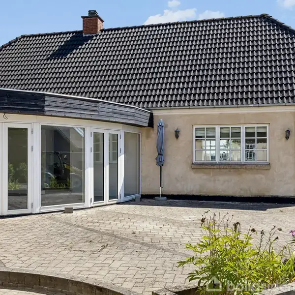 Hus med sort tegltag og beige facade; store vinduer i udestuen. Står på flisebelagt terrasse med nogle få blomster og planter. Blå paraply ved vindue. Klar himmel.