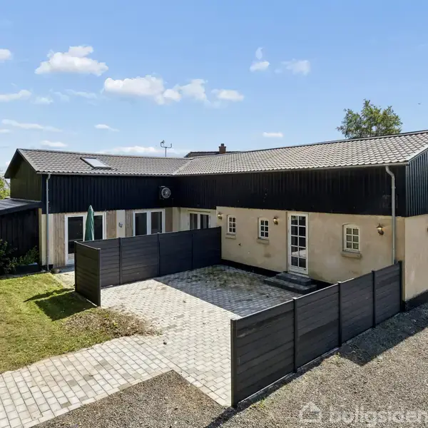Et hus med mørkt træværk og beige vægge har en indhegnet terrasse. Omkringliggende omgivelser inkluderer grusvej og græsareal, under en klar blå himmel med hvide skyer.