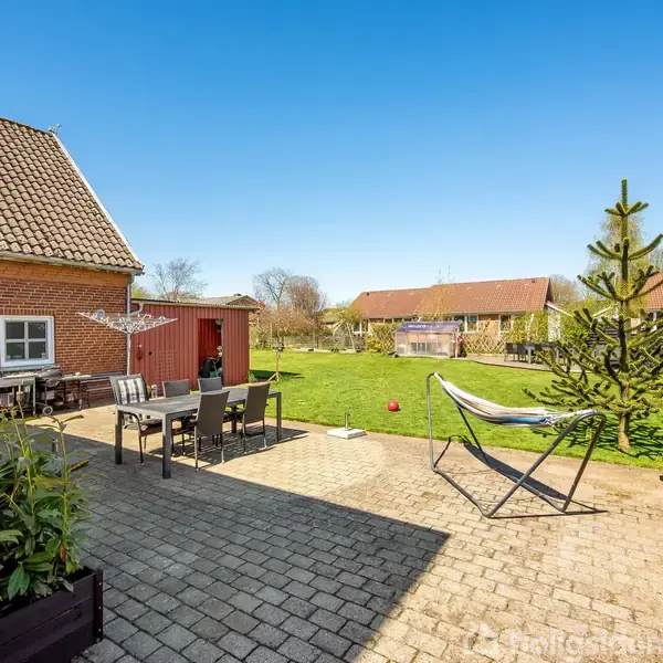 Terrasse med havemøbler placeret på stenterrasse ved siden af et rødt murstenshus. I baggrunden er en stor græsplæne, en hængekøje og flere mindre bygninger under en klar blå himmel.