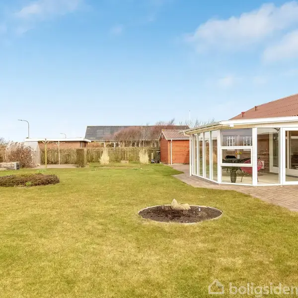 En solrig have med grøn græsplæne, flagstang og plantebede foran et rødt teglhustak og en lukket veranda. En hvid garage er synlig i baggrunden, omgivet af hække.