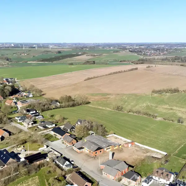 Landsby med spredte huse og gårde. Landskabet består af marker og bakker med træer, og en by ses i horisonten. Det er en solrig dag med klar, blå himmel.