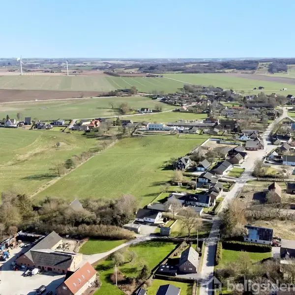 Landsbyen er omgivet af marker og spredte bygninger. Huse og gårde ligger langs veje, mens åbne marker strækker sig mod horisonten med et klart, solrigt vejr.
