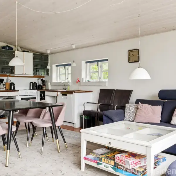 Sofa og sofabord med spil i en stue. Ved siden af står et spisebord med seks lyserøde stole. I baggrunden ses et køkken med hvide skabe og vinduer.