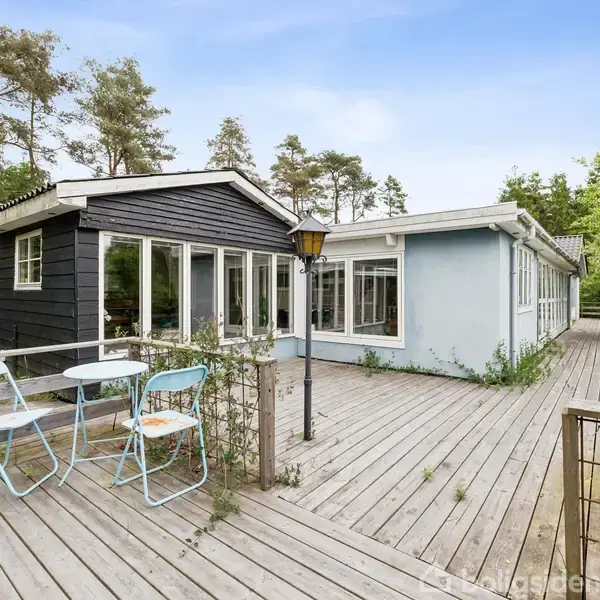 Sommerhus med sort facade, store vinduer og træterrasse med blå havemøbler. Beliggende i grøn, skovklædt område med lamper og en afslappende atmosfære.