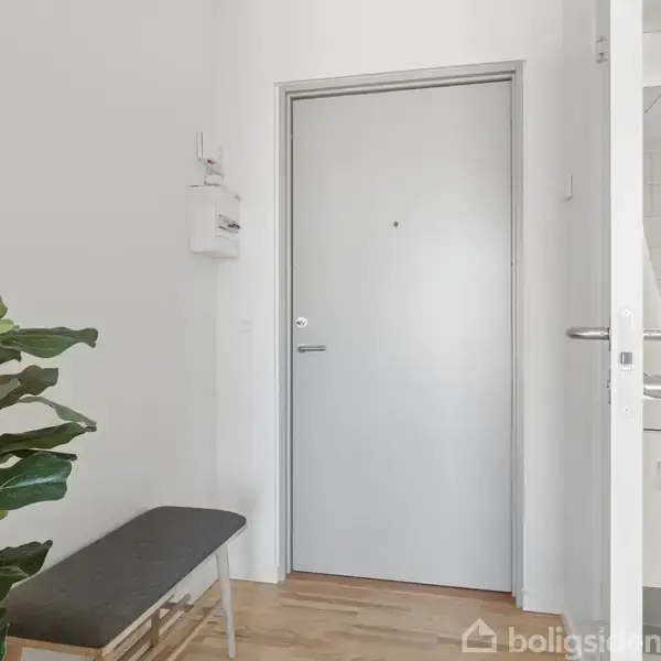 En grå dør står lukket i en lille entré med et plante på en bænk ved siden af, og en delvist åben dør fører ind til et flisebelagt rum.