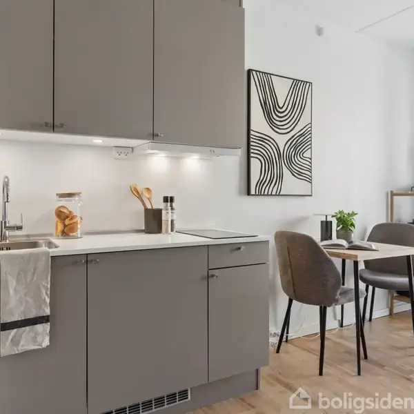 Grå køkkenskabe med hvid bordplade og håndvask. Ved siden af et lille spisebord med to stole. Vægdekoration og reol i baggrunden skaber moderne, minimalistisk atmosfære.