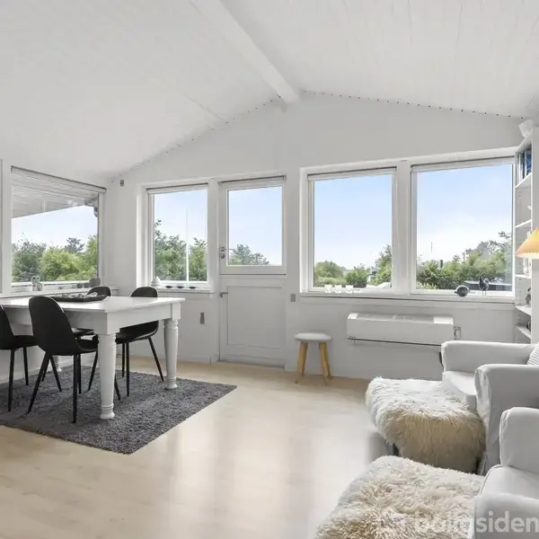 Et lyst, moderne opholdsrum med hvide vægge, indeholder et spisebord med fire sorte stole. Store vinduer lukker naturligt lys ind. En sofa og lænestol står til højre, dekoreret med tæpper.
