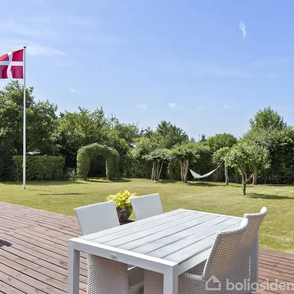 Et hvidt bord med stole står på en træterrasse. En flagstang med Dannebrog vimpler i haven, der er omgivet af buske. En hængekøje hænger mellem to træer.