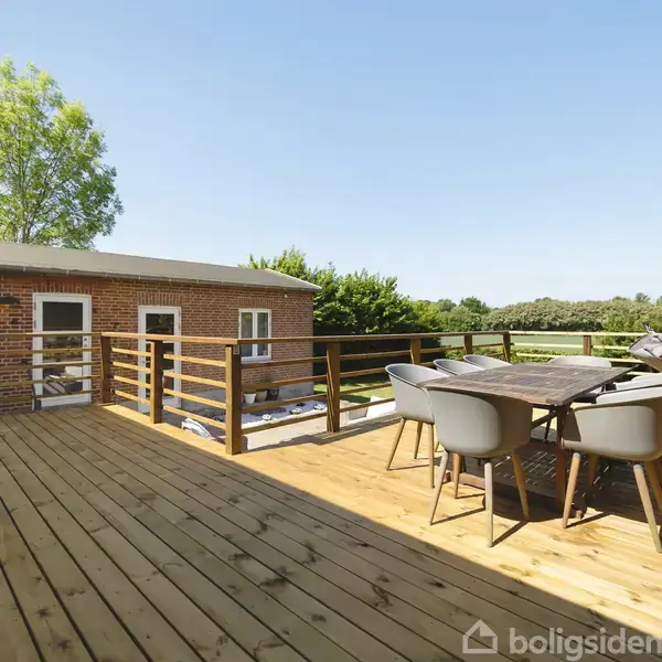 Terrasse med træmøbler i et landligt område. Terrassebord omgivet af stole. Baggrund indeholder en lys murstensbygning, grønne træer og mark, og himlen er klar og blå.