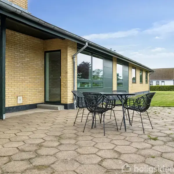 En flisebelagt terrasse med et sort metalbord og fire stole står ved siden af et gult murstenshus med store vinduer. Græsplæne og himmel udgør baggrunden.