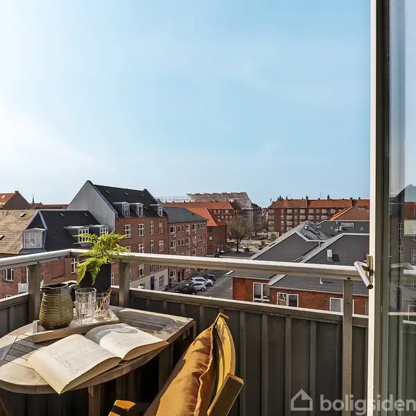 Et lille rundt havebord med en åben bog og en planter i en potte står på en altan. Man ser en udsigt over en by med rødstensbygninger under en klar himmel.