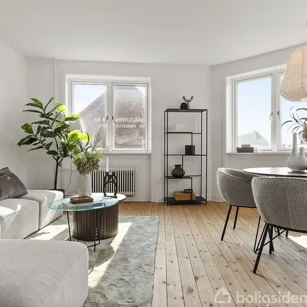 En lys stue med moderne indretning indeholder en hvid sofa, rundt sofabord, grå stole ved et spisebord og reol. Vinduerne slipper sollys ind. Tekst: "ÅBENT VANGEGÅRD 4 NOVEMBER."
