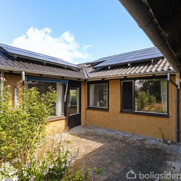 Et enkelt etplanshus med solpaneler på taget. Huset har gule mursten og store vinduer. Foran er der en gårdhave med fliser og nogle buske i dagtimerne.