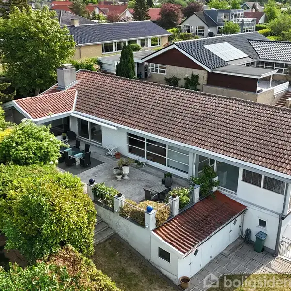 Rødt tegltagshus med stor terrasse omgivet af frodige haver. Huset er beliggende i et boligområde med flere lignende huse og har solpaneler synlige på naboens tag.