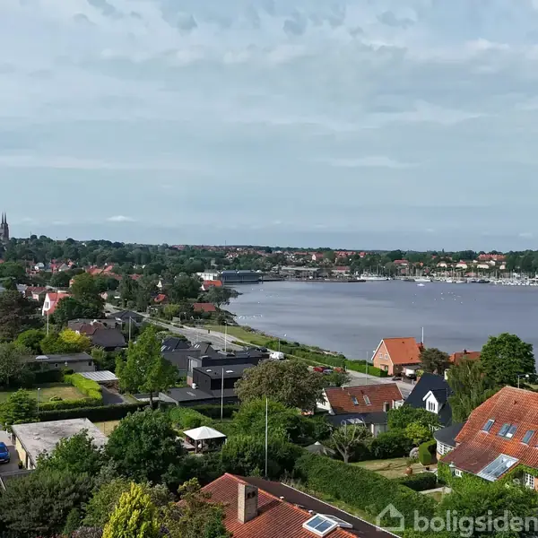 Udsigt over et kvarter med rødt tagede huse, der strækker sig mod en bugt, med en kirke på en bakketop i det fjerne, omgivet af grønne træer og græsarealer.