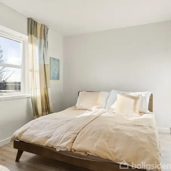 En dobbeltseng står centralt i et lyst soveværelse. Sengen er redt med lyse beige dyner og puder. Til venstre ses et vindue med gardiner og en grøn plante.