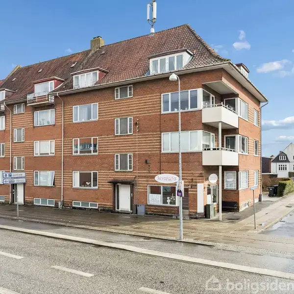 En murstensbygning med rødt tag har flere altaner og vinduer. Bygningen står ved en asfalteret vej med fortov. Skiltet på facaden viser teksten: “Cafe Victoria Pizza Kebab House”.