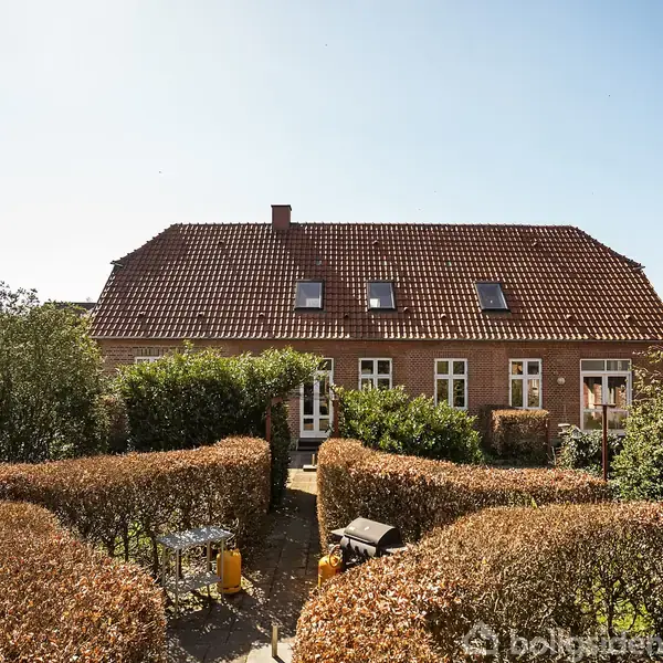 Huset står stille, omgivet af en velplejet have med formklippede buske og en terrasse. Rød murstensfacade med rødt tegltag og kviste. Klar blå himmel bagved.