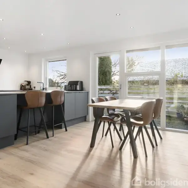 Et moderne køkken med en køkkenø og barstole, omgivet af spisebord med stole. Store vinduer giver udsigt til en have med træer og en grill. Ingen tekst i billedet.