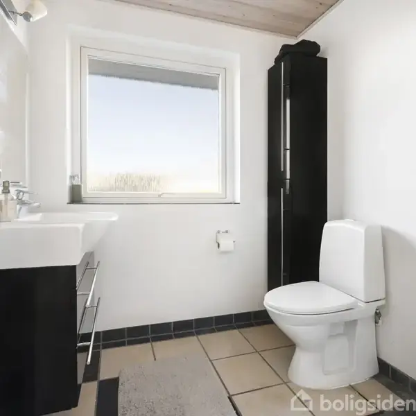 Et badeværelse med et toilet står ved siden af en høj, sort skab. Håndvasken er monteret til venstre. Et vindue giver naturligt lys ind i rummet.