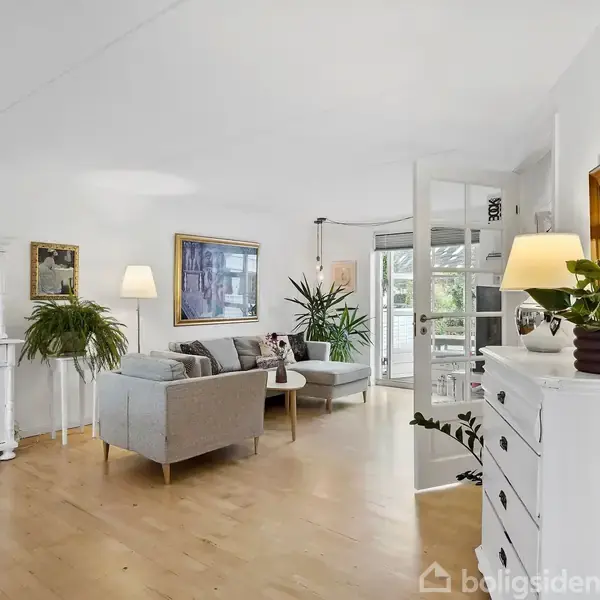 Stue med grå sofaer står i et lyst rum med trægulv. Møbler og planter dekorerer rummet, mens store vinduer bagtil bringer lys ind. Rundt spredte lamper giver varm belysning.