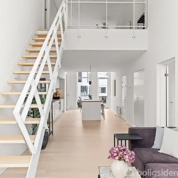 En moderne stue med lofttrappe til venstre leder opad. Midt i rummet står et spisebord med stole. Hvide vægge og store vinduer giver lys i rummet.