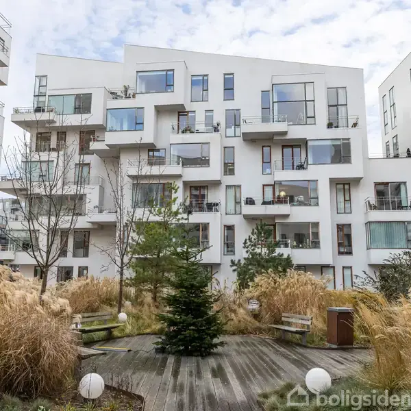 Lejlighedskompleks med moderne, hvide facader. Bygningen har flere balkoner og store vinduer. I forgrunden ses en gårdhave med træer, buske og bænke, omgivet af høje græsplanter.