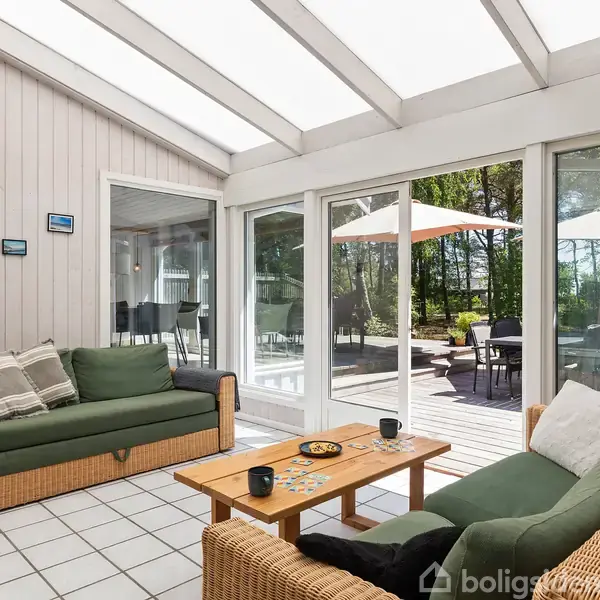 To flettede sofaer med grønne hynder står i en lys stue med glasvægge. Sofabordet mellem dem har kopper og brætspil. Udenfor ses en terrasse med parasoller og skov.
