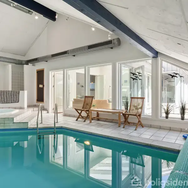En indendørs swimmingpool omgivet af flisegulv, med bord og stole ved siden af store vinduer. Blå loftsbjælker og dekorative planter i hjørnet fuldender den lyse, afslappende atmosfære.