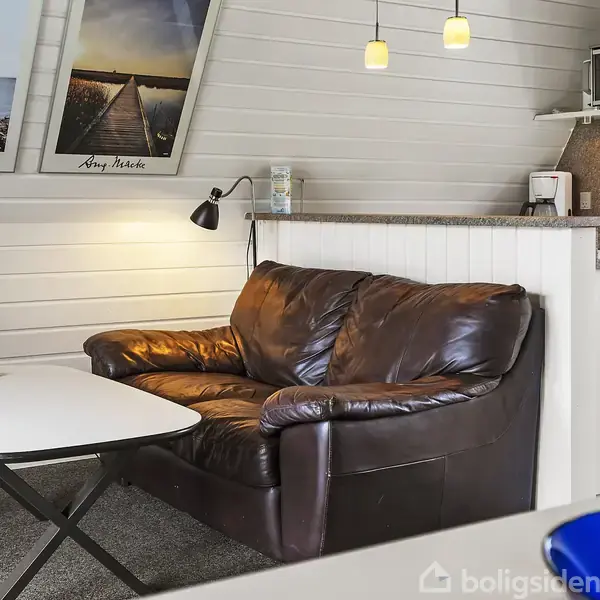 Brun lædersofa står ved siden af et hvidt bord med fjernbetjening; hængelamper lyser rummet op. Køkkenet med vask og bordplader ses i baggrunden. Væggene er dekoreret med billeder.
