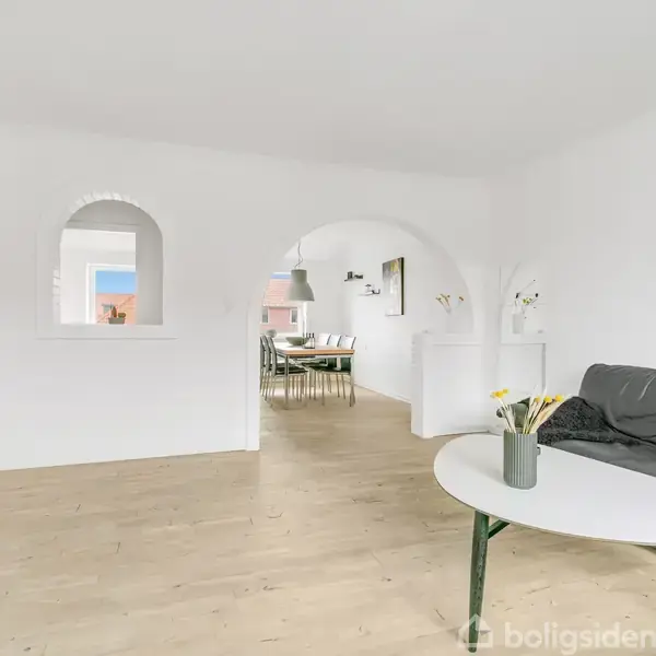 Sofa i en stue med åben buegang, står ved et ovalt bord med vase. Lys træetage flankeret af dekorationsgenstande og et verdenskort på væggen.