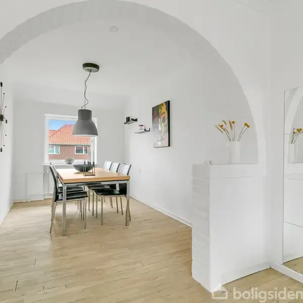Et spisebord med stole står midt i et lyst rum. En pendellampe hænger over bordet. Der er en hvidbuet væg og et spejl, som reflekterer interiøret.