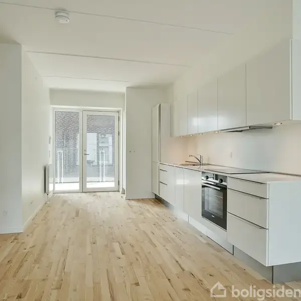 Køkken med hvide skabe og indbygget ovn langs en væg. Trægulv strækker sig gennem rummet mod glasdøre, der giver udsigt til en balkon.