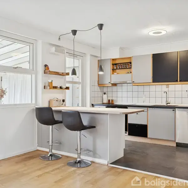 Et moderne køkken med to sorte barstole ved en hvid ø. Sort-hvide skabe og flisevæg. Lamper hænger over øen. Vindue og hylder på venstre væg.