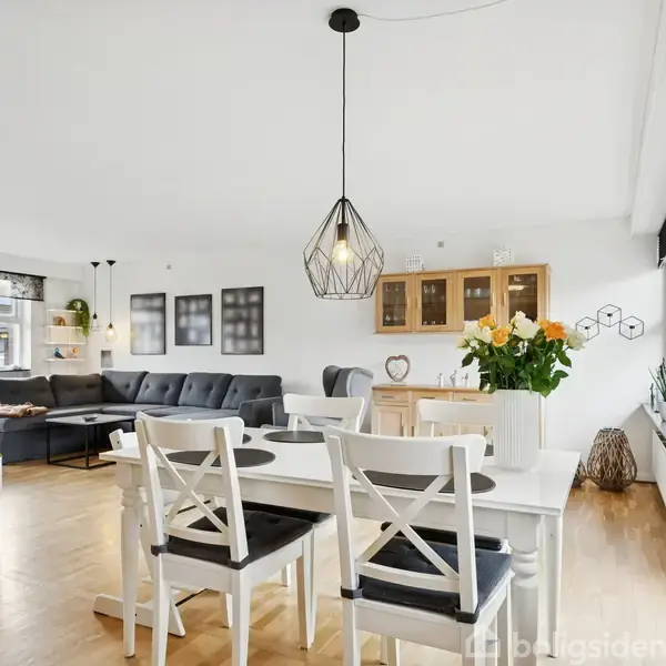 En lys spisestue indeholder et hvidt bord med fire stole, beliggende foran store vinduer med udsigt. Til venstre ses en grå sofa, vægge med kunst og grønne planter.