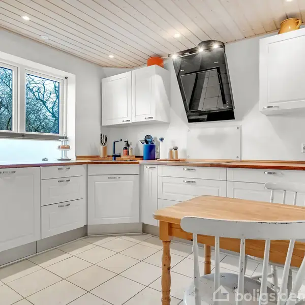 Et lyst, moderne køkken med hvide skabe og en sort emhætte. Et lille træbord med en stol står centralt. Store vinduer giver udsigt til træer udenfor.