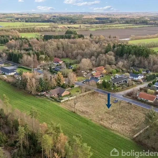 Luftfoto viser en mark med en blå pil, placeret ved en landevej. Omgivet af huse, skov og åbne marker i landlig omgivelser.