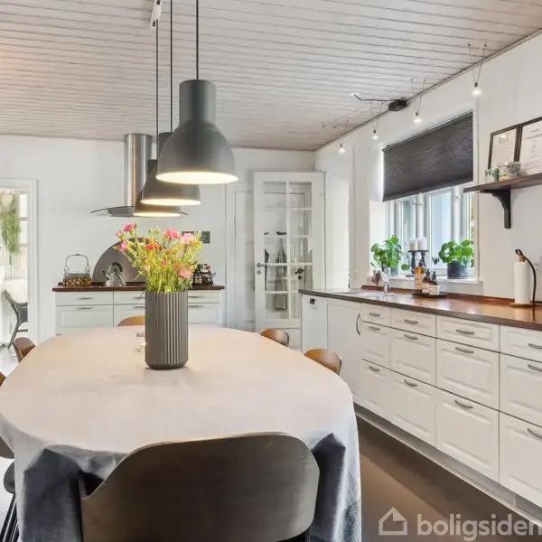 Et lyst køkken med et langt spisebord dækket med en dug og blomster i en vase. Ved siden af er hvide skabe og en bordplade med planter og en kaffemaskine.