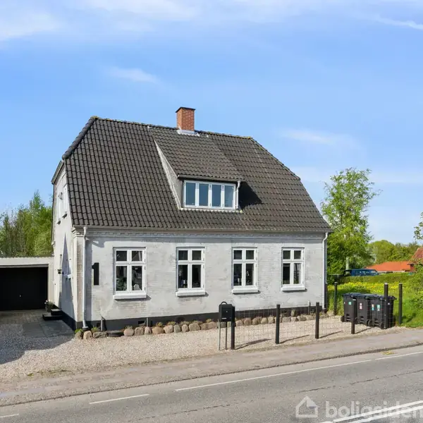 Et hvidt hus med sort tag står ved en vej. Til venstre er en garage og en parkeret bil. Baggrunden rummer grønt løv og en klar himmel.