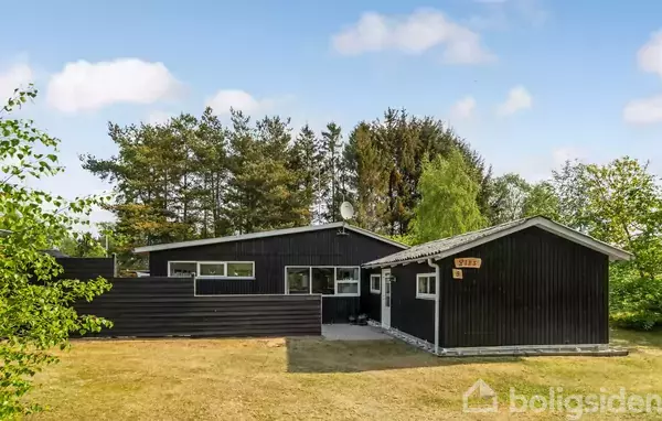 Et sort træhus står stille omgivet af grønne træer og buske. Baggrunden består af en lys blå himmel med få skyer.