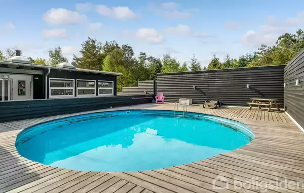 Rund swimmingpool med klart blåt vand, omgivet af en træterrasse. I baggrunden ses en bygning med vinduer, træbænke og en lyserød stol. Beliggende i et grønt, skovklædt område.