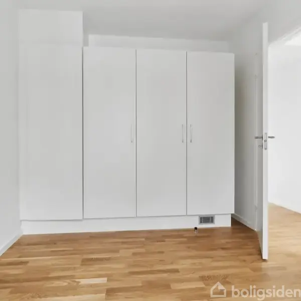 Et hvidt skab står mod en væg; det er lukket, og rummet har trægulv og hvide vægge med en åben dør til en tilstødende lys gang.