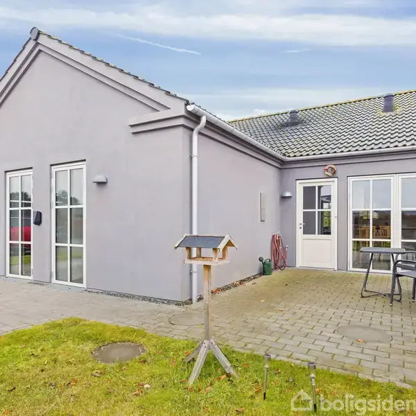 Gråt enfamiliehus står stille på en terrasse. Bord og stole står ved siden af huset. Græsplæne og fuglefoderbræt fylder forhaven.