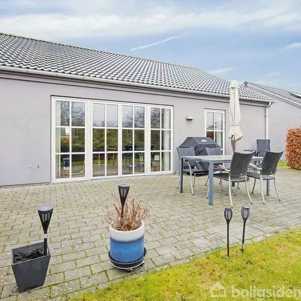 En terrasse med havemøbler og en grill står på flisebelægningen foran et moderne hus med store vinduer. Baghaven er omgivet af hække og græsplæne.