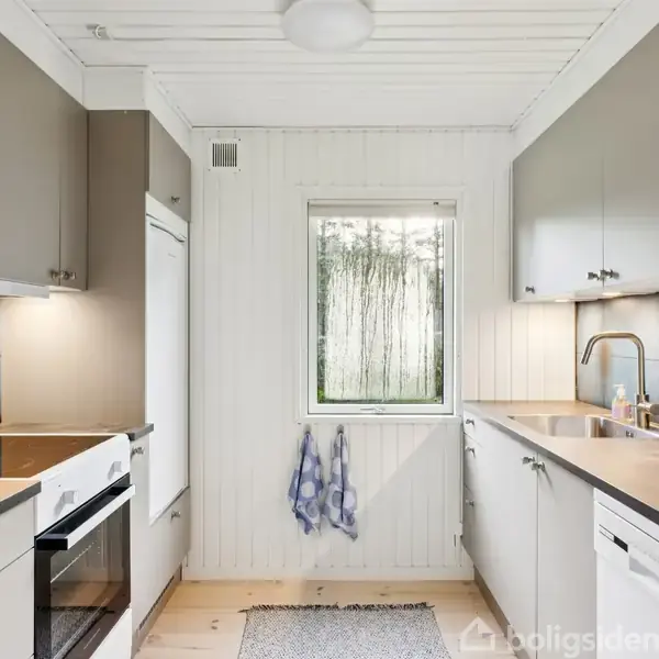 Et moderne køkken med grå skabe og træbordplade. En ovn med komfur står til venstre. Der er en vask til højre. Et vindue med udsigt og et blåhvidt håndklæde hænger under det.