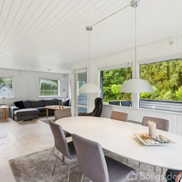 Et moderne, lyst rum med et ovalt spisebord omgivet af stole. Store vinduer slipper naturligt lys ind, og sofaen står ved vinduerne. Grønne træer ses udenfor.