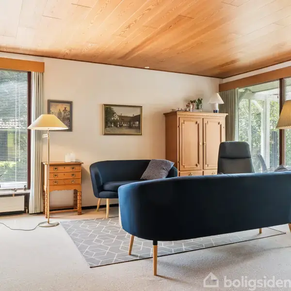 Møbleret stue med to mørkeblå sofaer på et gråt tæppe. Store vinduer giver udsigt til en grøn have. Rummelige reoler, aflange billeder og lamper skaber en hyggelig atmosfære.