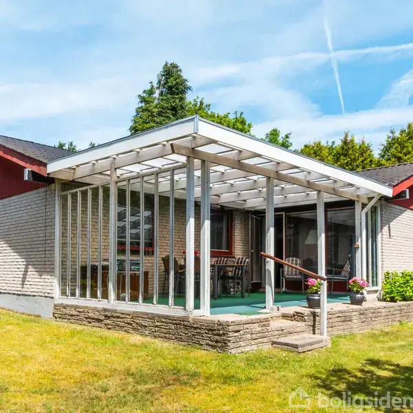 Et parcelhus i én etage med en stor glasoverdækket terrasse. Huset er omgivet af en velholdt græsplæne og buske. Himmel med lette skyer.